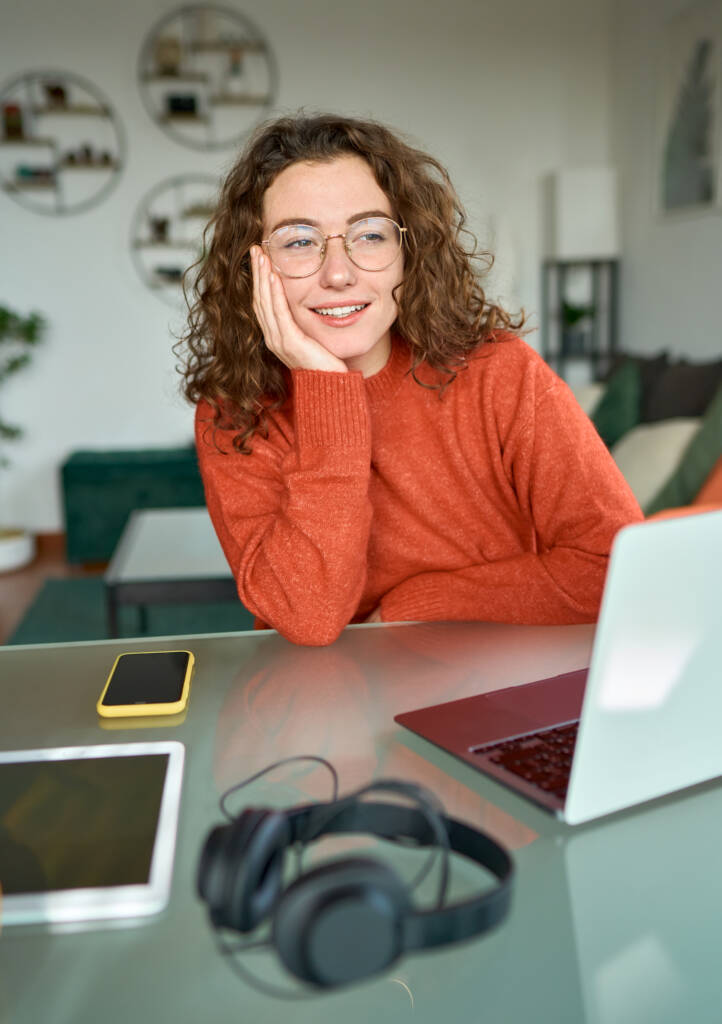 Young happy woman looking away and thinking while working on laptop at home.